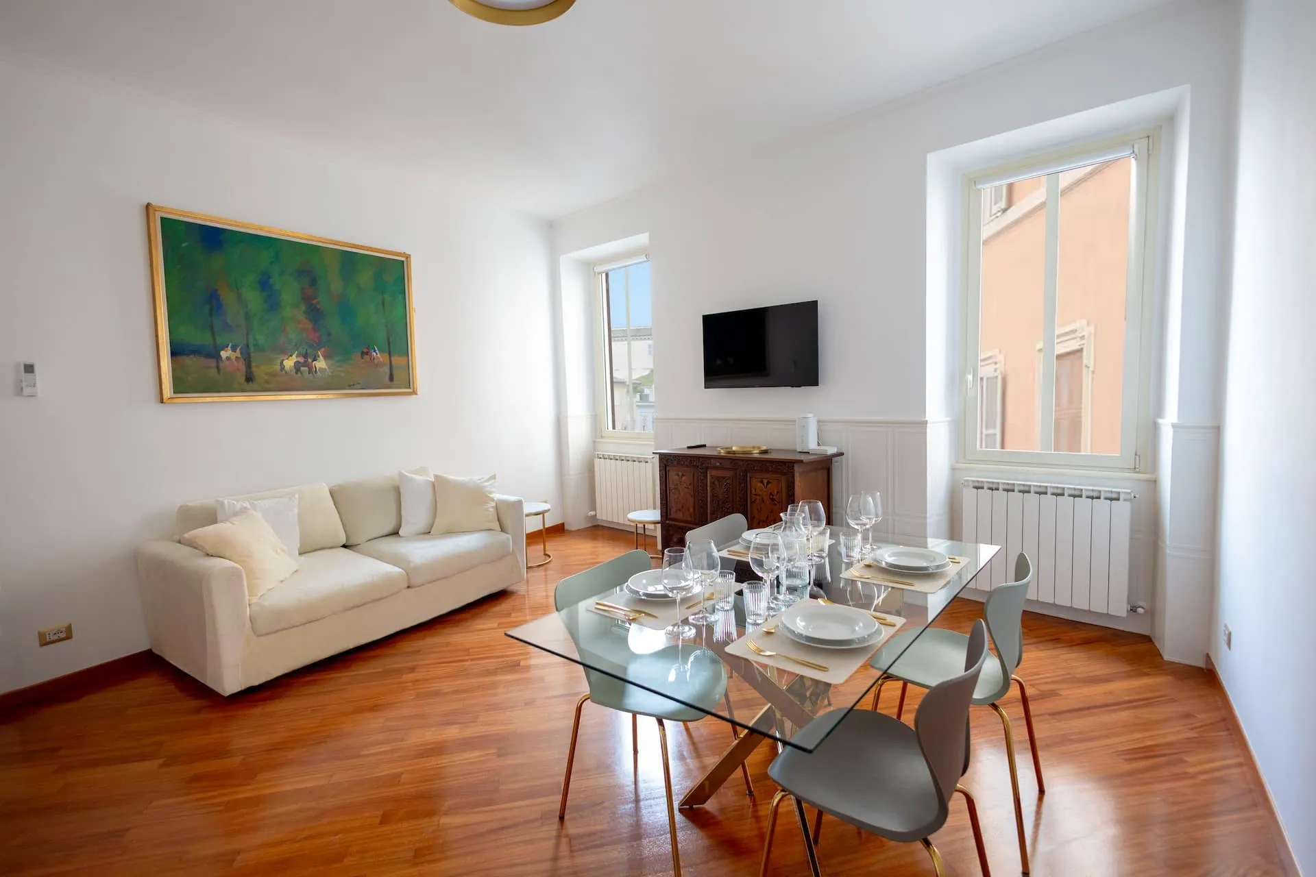 Soggiorno luminoso con parquet, divano bianco, tavolo di vetro apparecchiato e un quadro verde alla parete