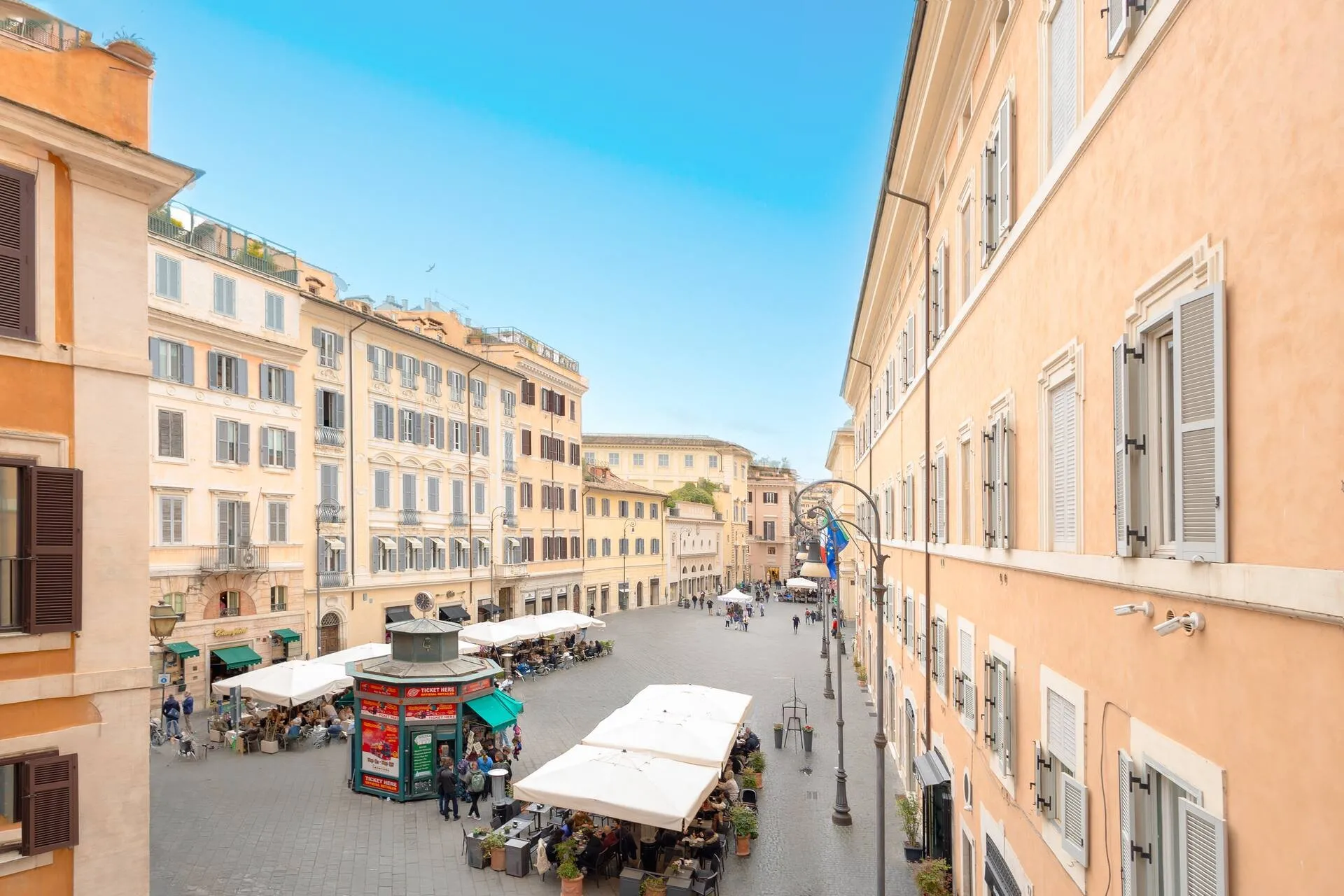 Vista dalla finestra sulla piazza, coi tavolini dei caffè e le facciate color pesca