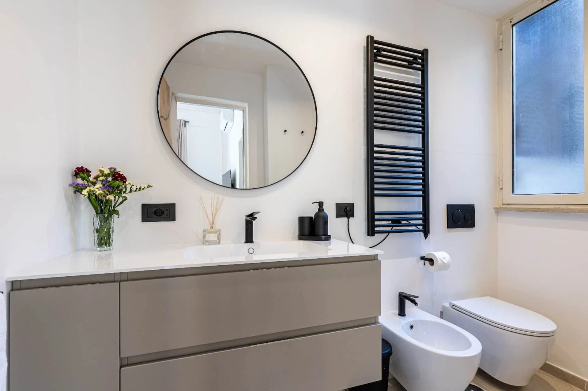 Bagno con mobile grigio, specchio rotondo e scaldasalviette nero