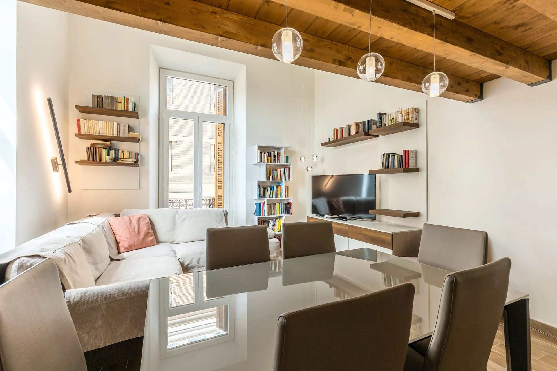 Zona pranzo con tavolo di vetro e sedie grigie, librerie sulle pareti e finestra sul fondo