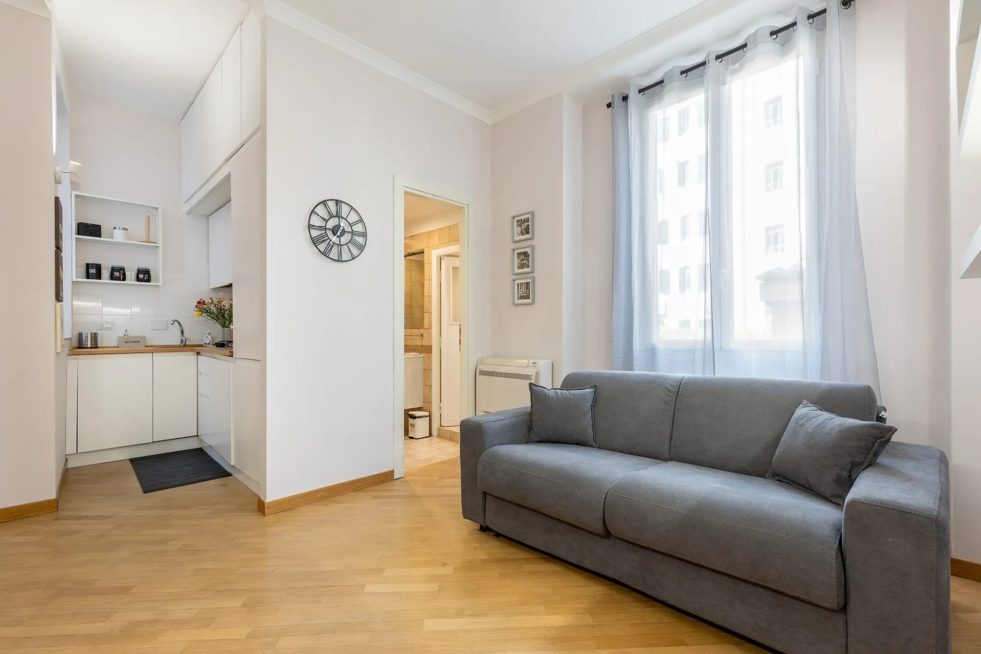 Soggiorno con divano grigio, parquet e finestra, cucina bianca sul fondo