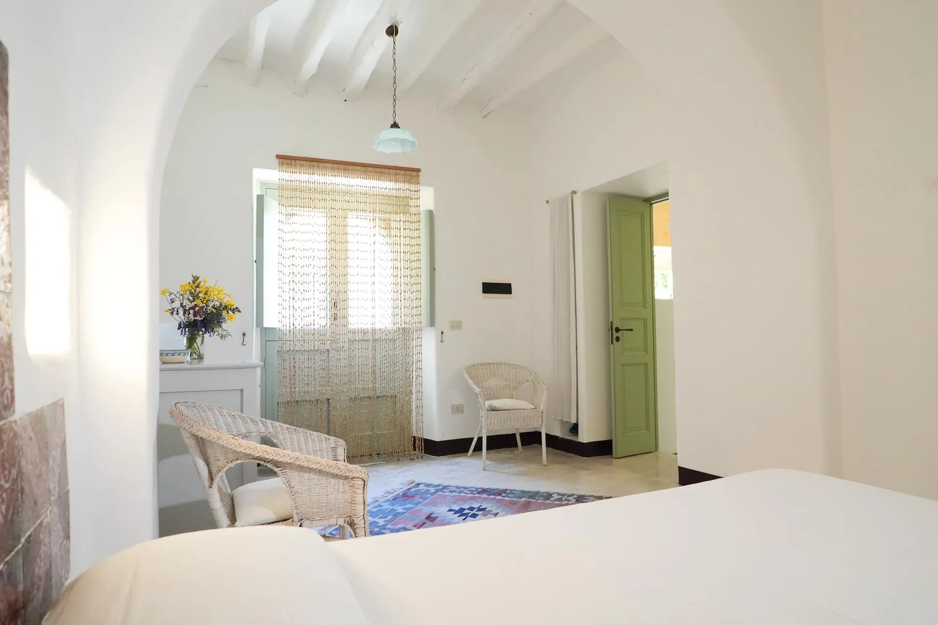 Camera bianca sotto un arco, con un letto, una poltrona di vimini e la porta finestra