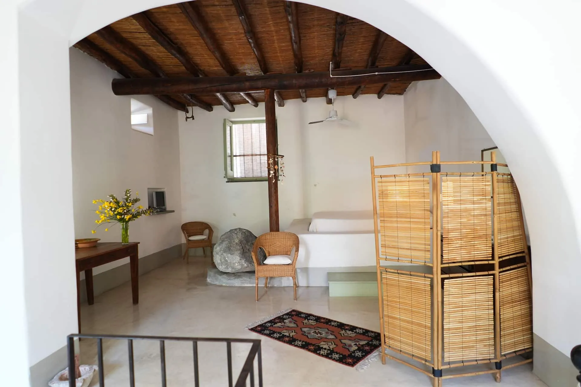 Stanza bianca sotto un arco con travi di legno, letto in muratura e paravento di vimini