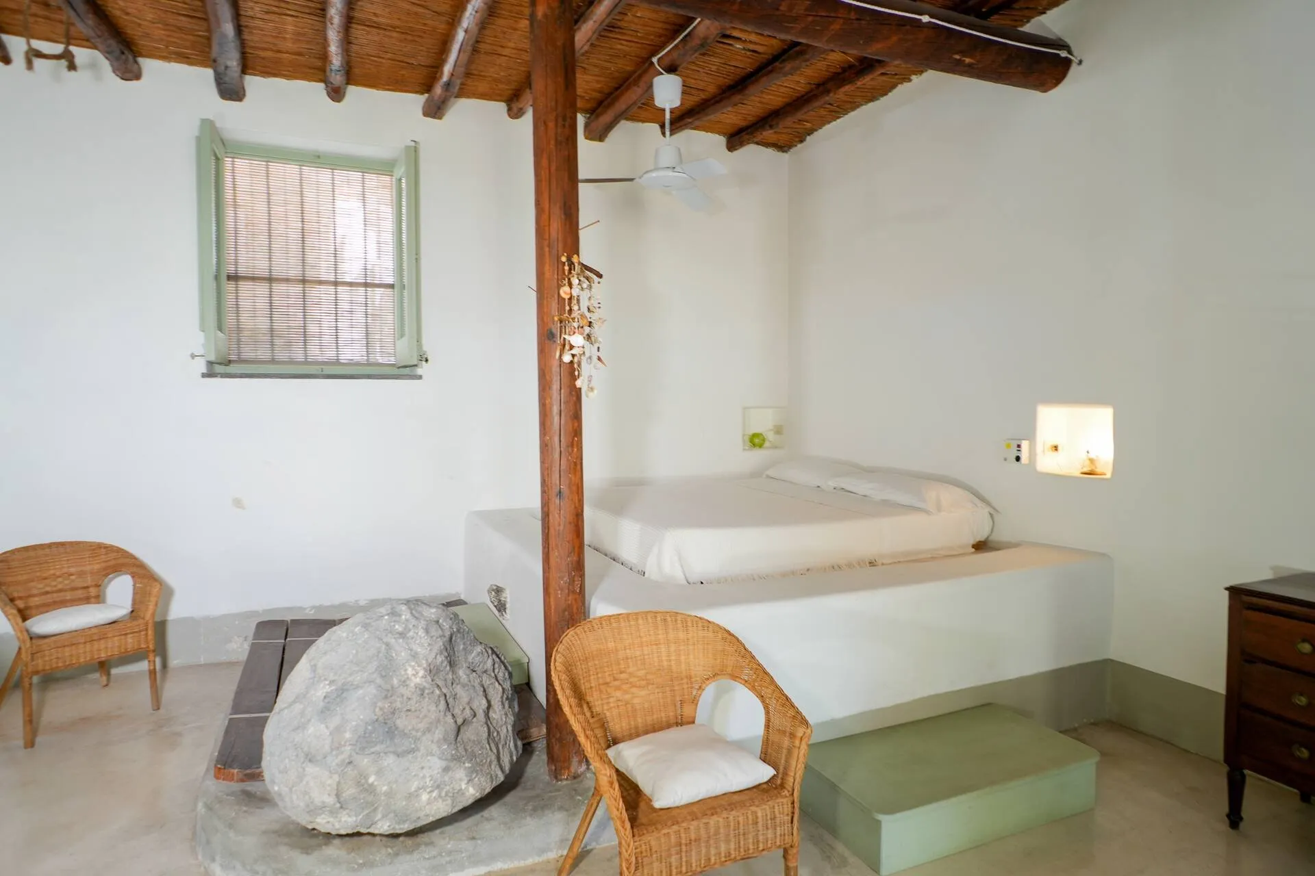 Letto in muratura, poltrone di vimini e un grande sasso, sotto il soffitto a travi