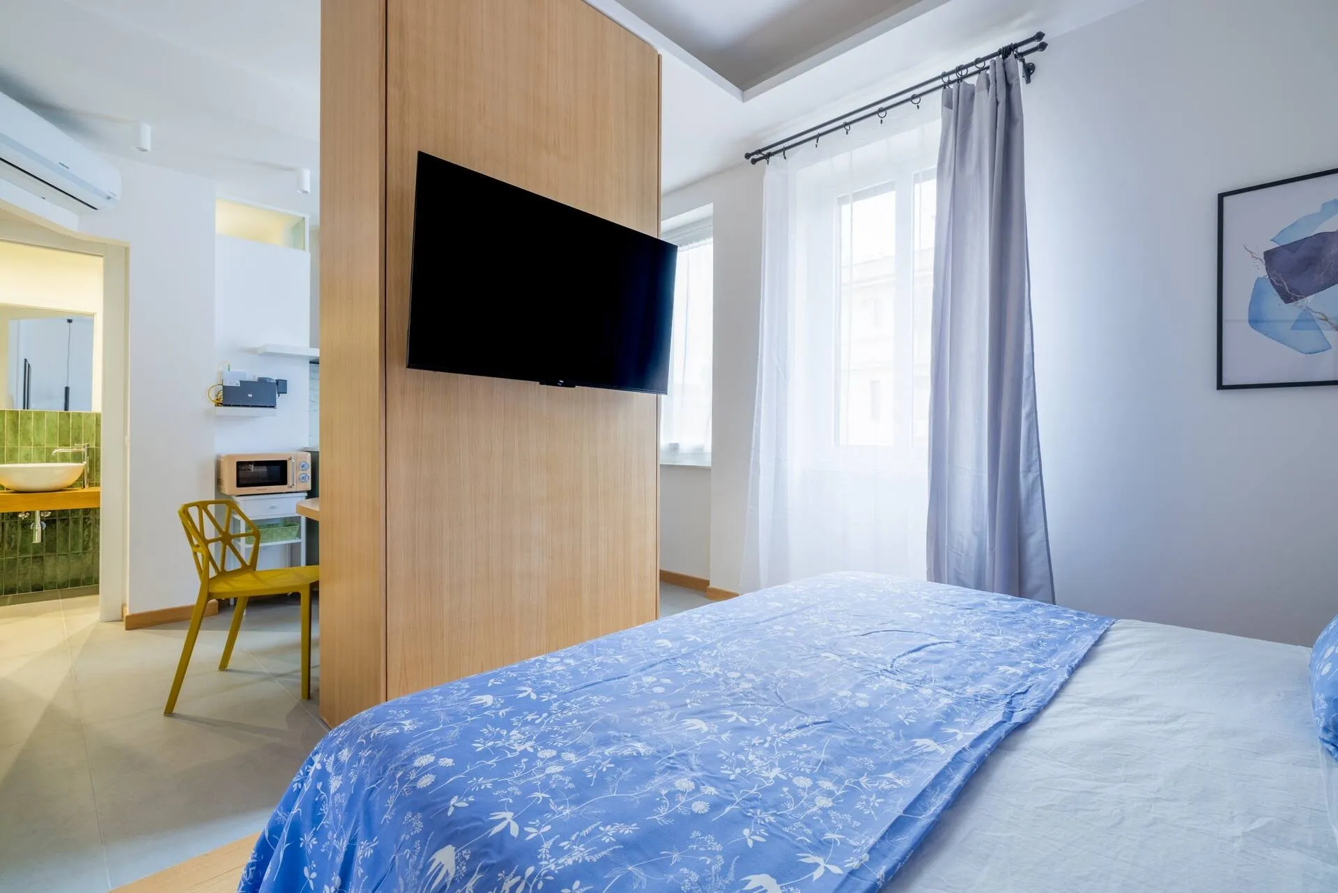 Letto matrimoniale e televisore sul divisorio di legno, finestra con tenda grigia