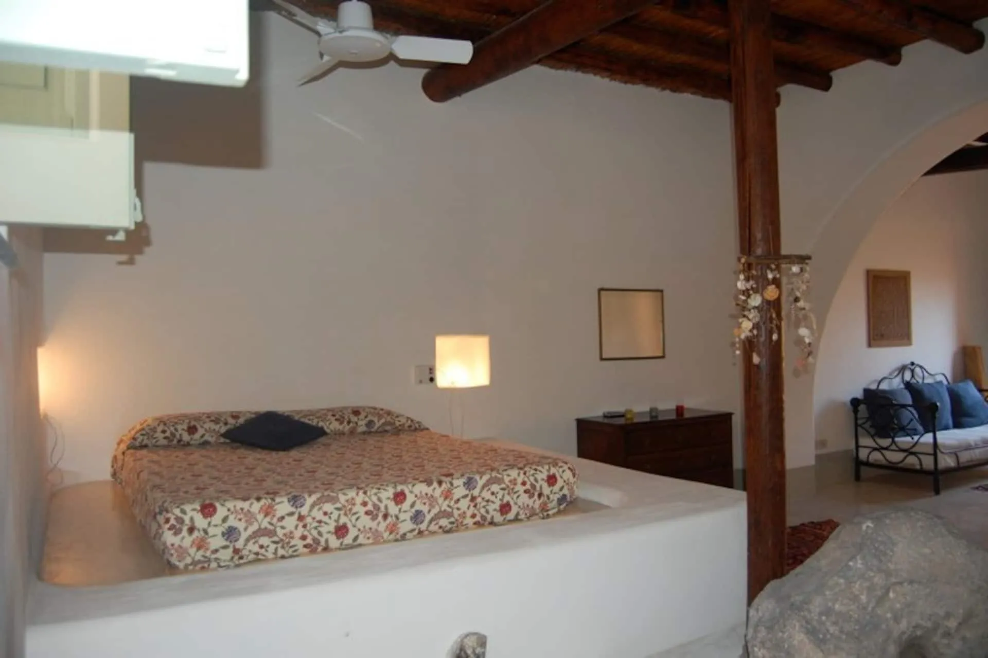 Letto matrimoniale in muratura sotto le travi di legno, con un divano in ferro battuto sul fondo