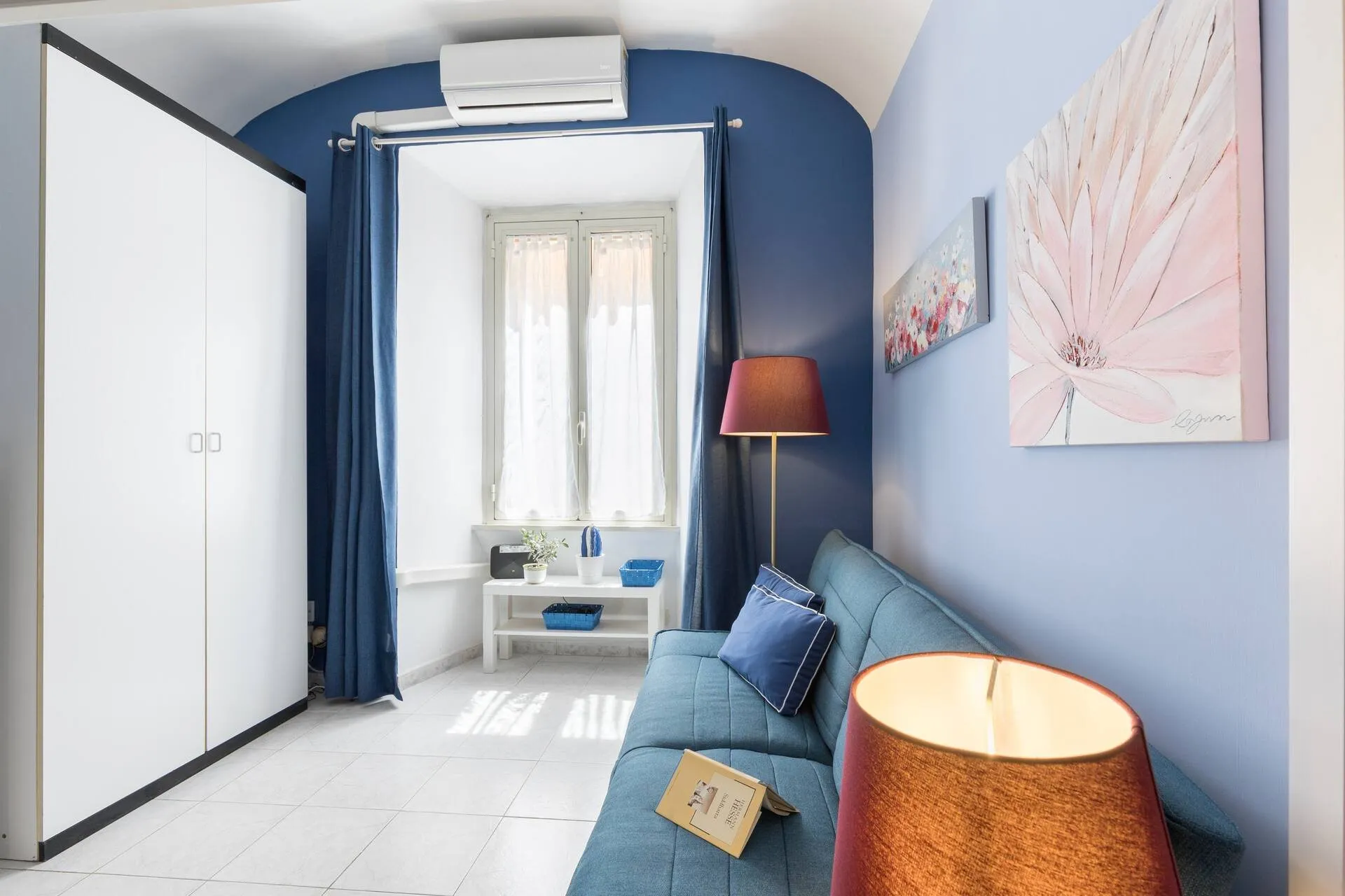 Divano blu, lampada e finestra con tende blu, armadio bianco a sinistra