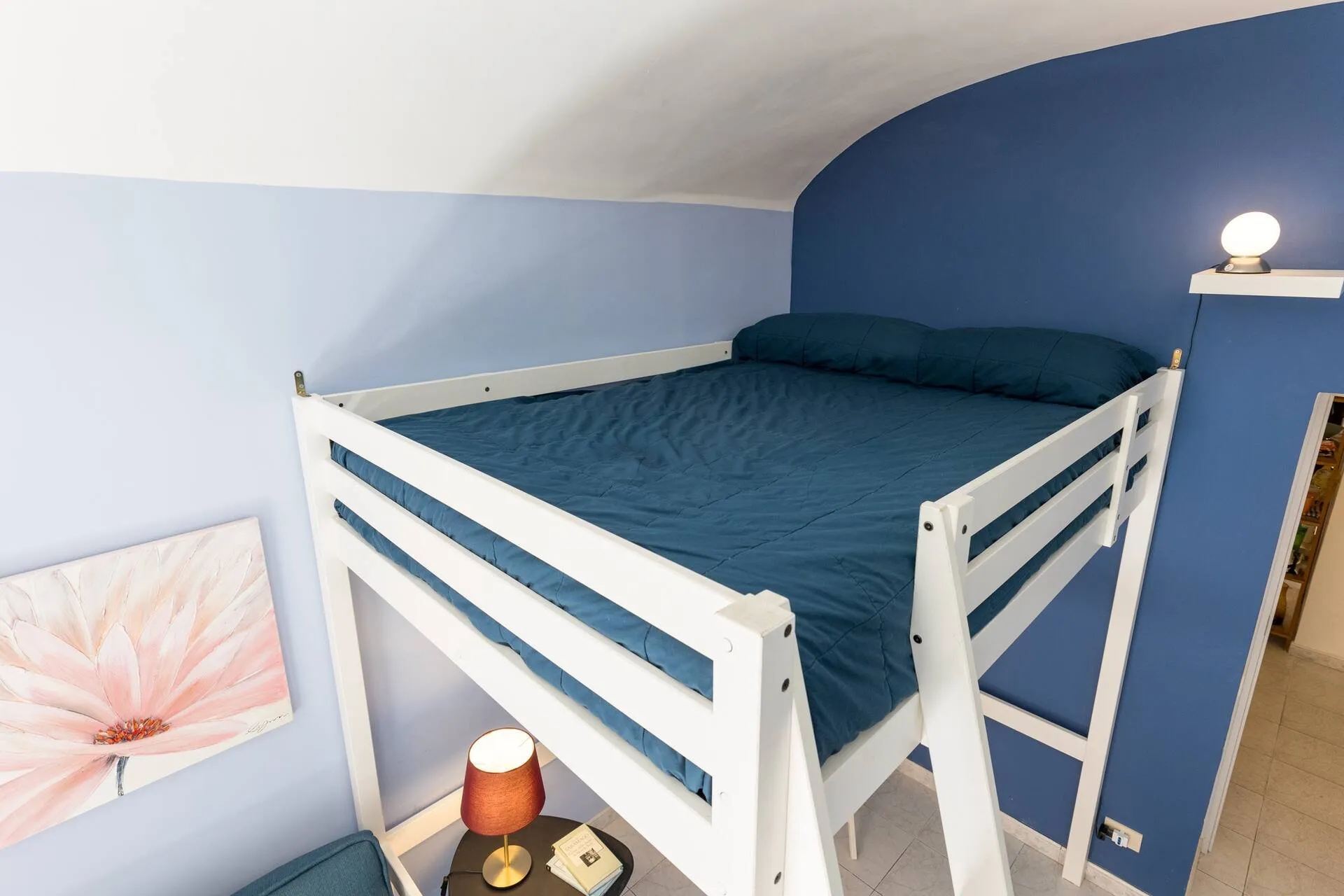 Letto matrimoniale sul soppalco bianco, parete blu e quadro di fiori