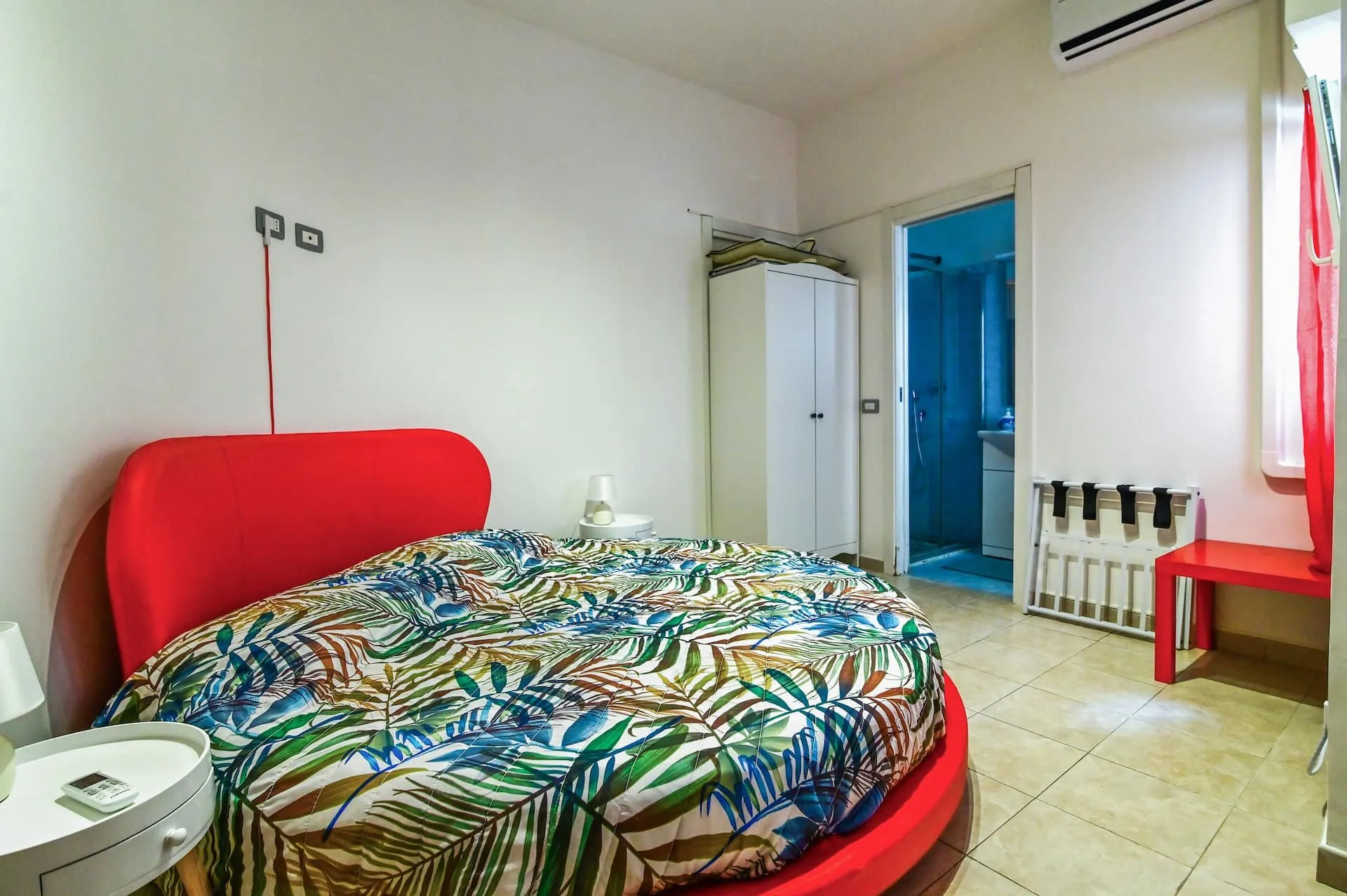 Letto rotondo con testiera rossa, armadio bianco e porta sul bagno