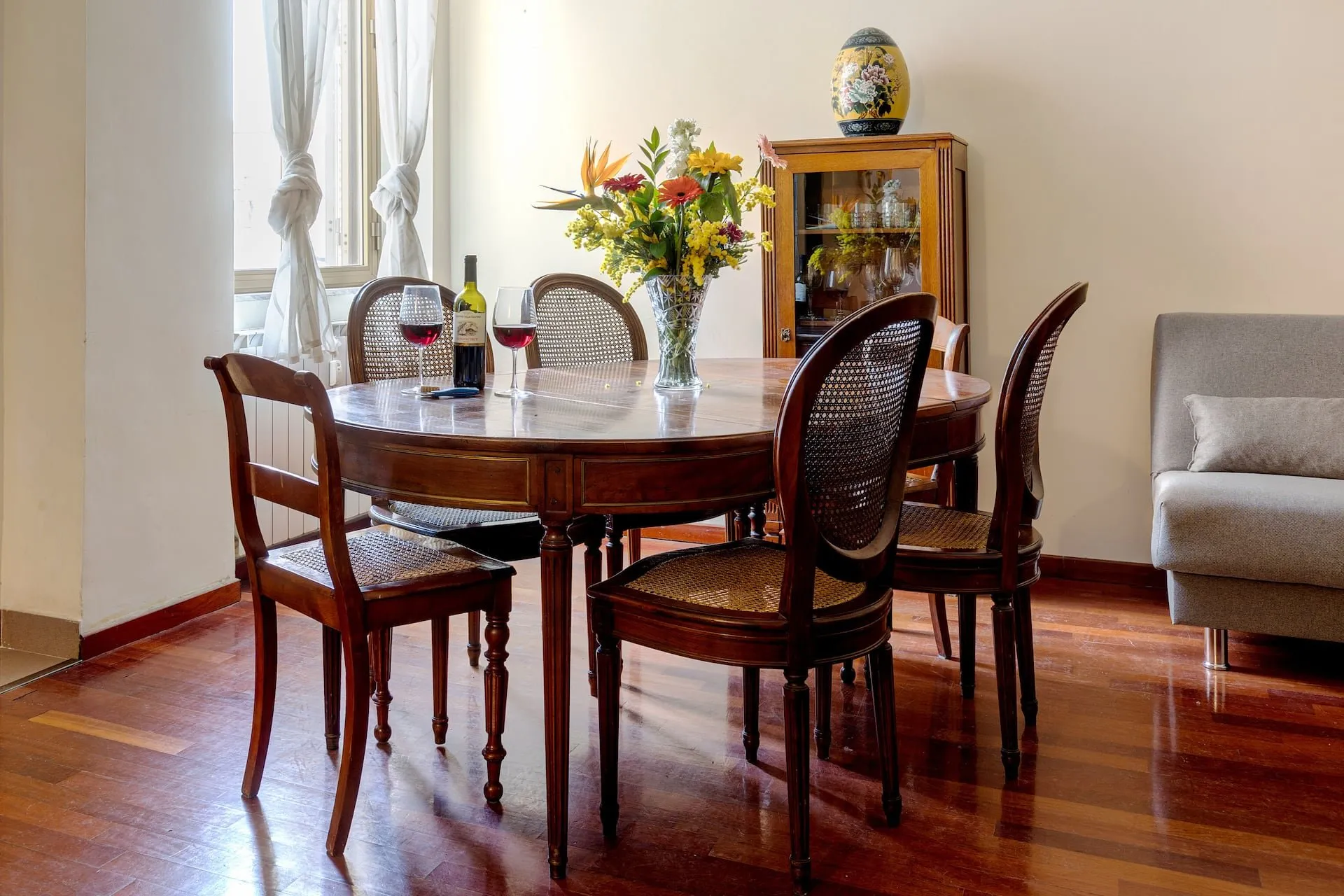 Tavolo da pranzo rotondo con sedie in legno scuro, vetrina e fiori, davanti alla finestra