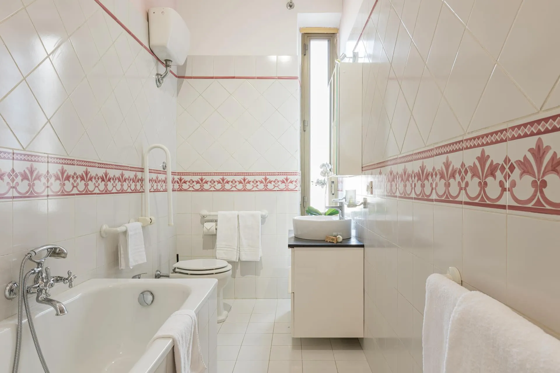 Bagno bianco con vasca, fregio rosso sulle piastrelle e finestra stretta